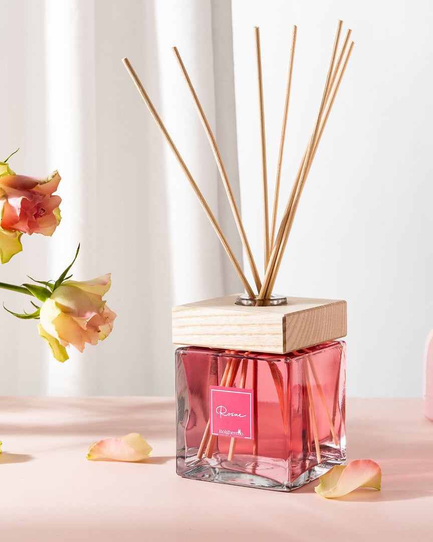 Diffusore di fragranze Bolgherello con liquido rosa e bastoncini di legno, affiancato da due rose e petali sparsi su una superficie rosa.